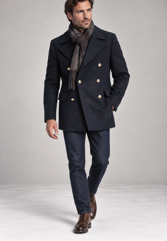 A man wearing a navy double-breasted wool peacoat with a scarf, dark jeans and brown boots